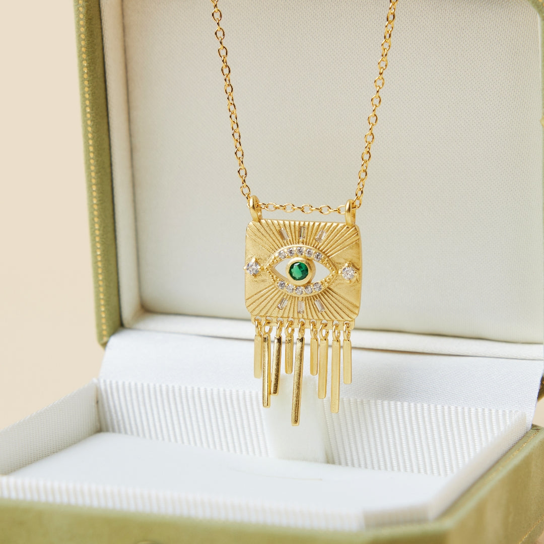 Collier doré avec un pendentif en forme d'œil stylisé, orné d'une émeraude centrale et de diamants, suspendu à une chaîne délicate.