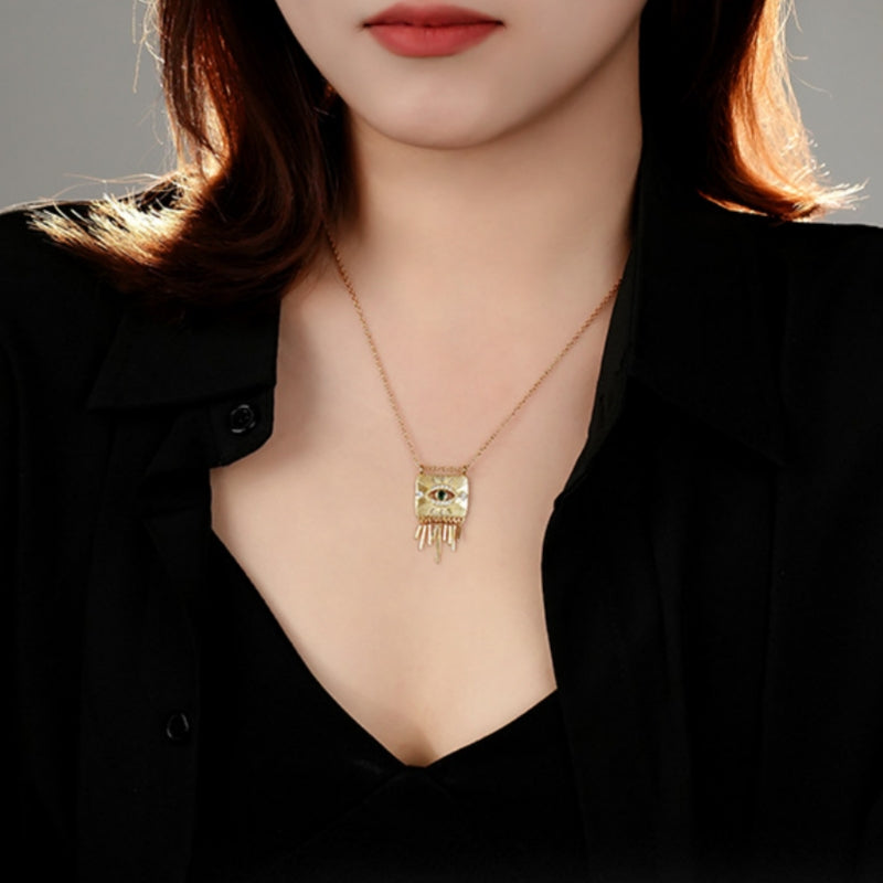 Collier plaqué or avec un pendentif carré orné d'un œil protecteur stylisé et de franges dorées.