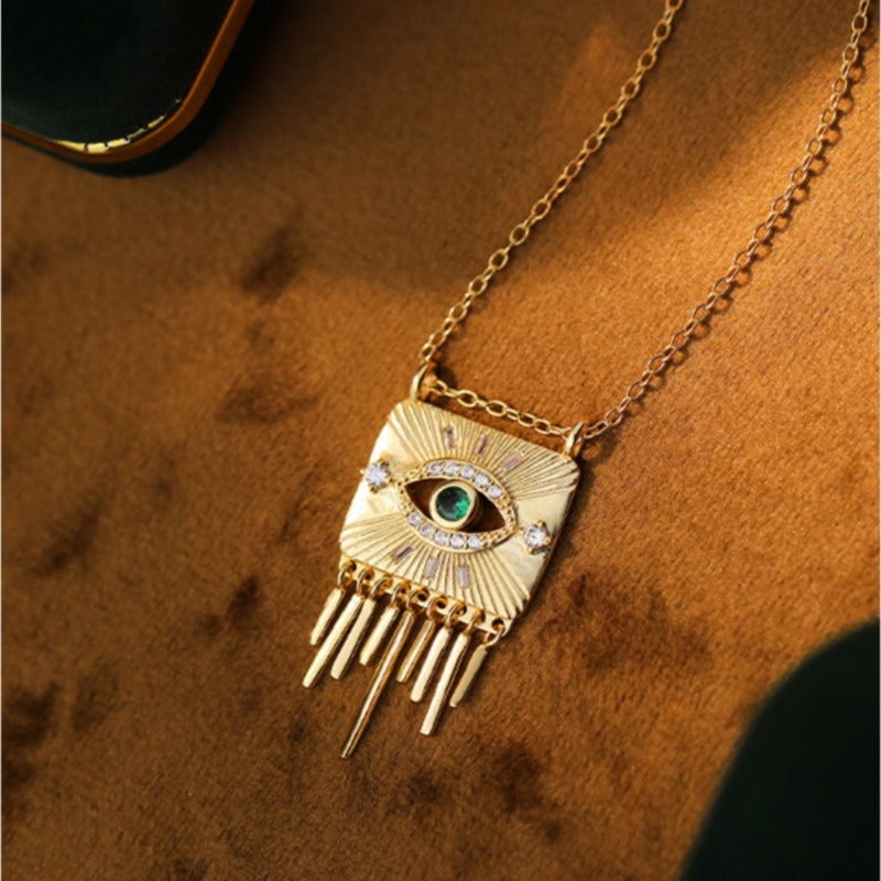 Collier plaqué or avec pendentif carré orné d'un œil stylisé serti d'une émeraude et de diamants, bordé de rayons et de franges tombantes.