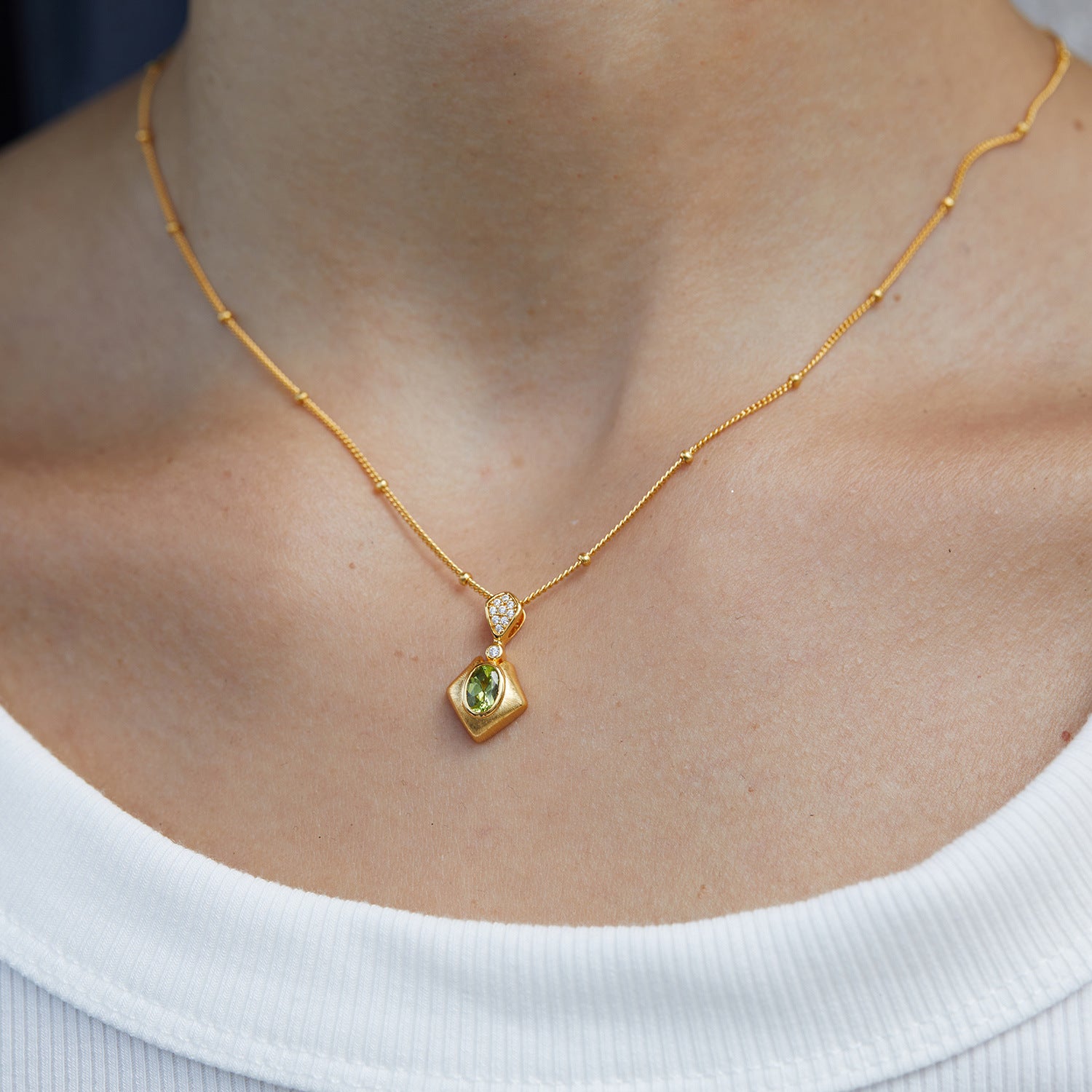 Gros plan sur un collier plaqué or avec un pendentif en forme de losange contenant une péridot verte et de petits diamants, porté par une femme sur un décolleté blanc.