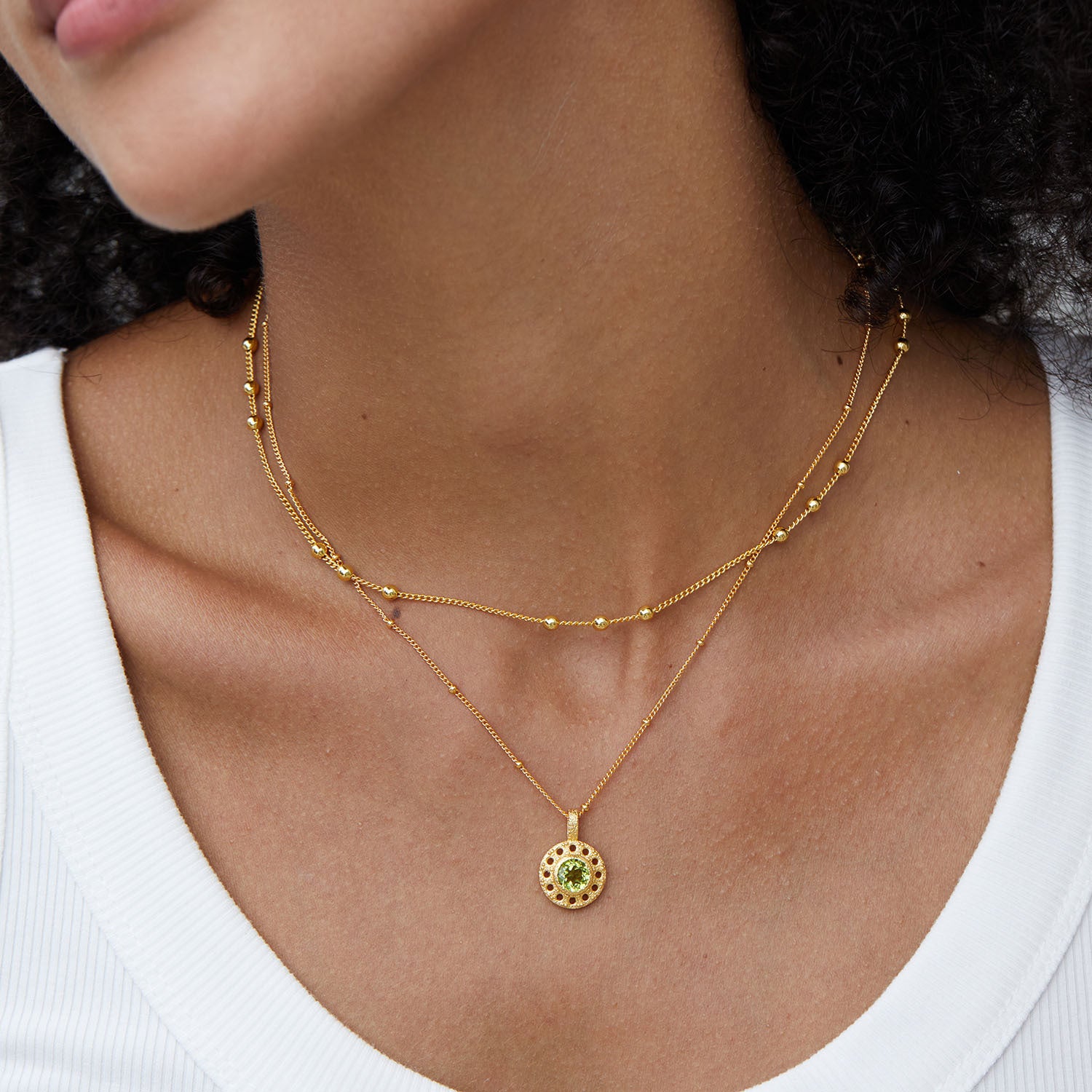 Gros plan sur un collier double chaîne en argent plaqué or, avec un pendentif rond orné d'une pierre de péridot verte au centre, porté par une femme.