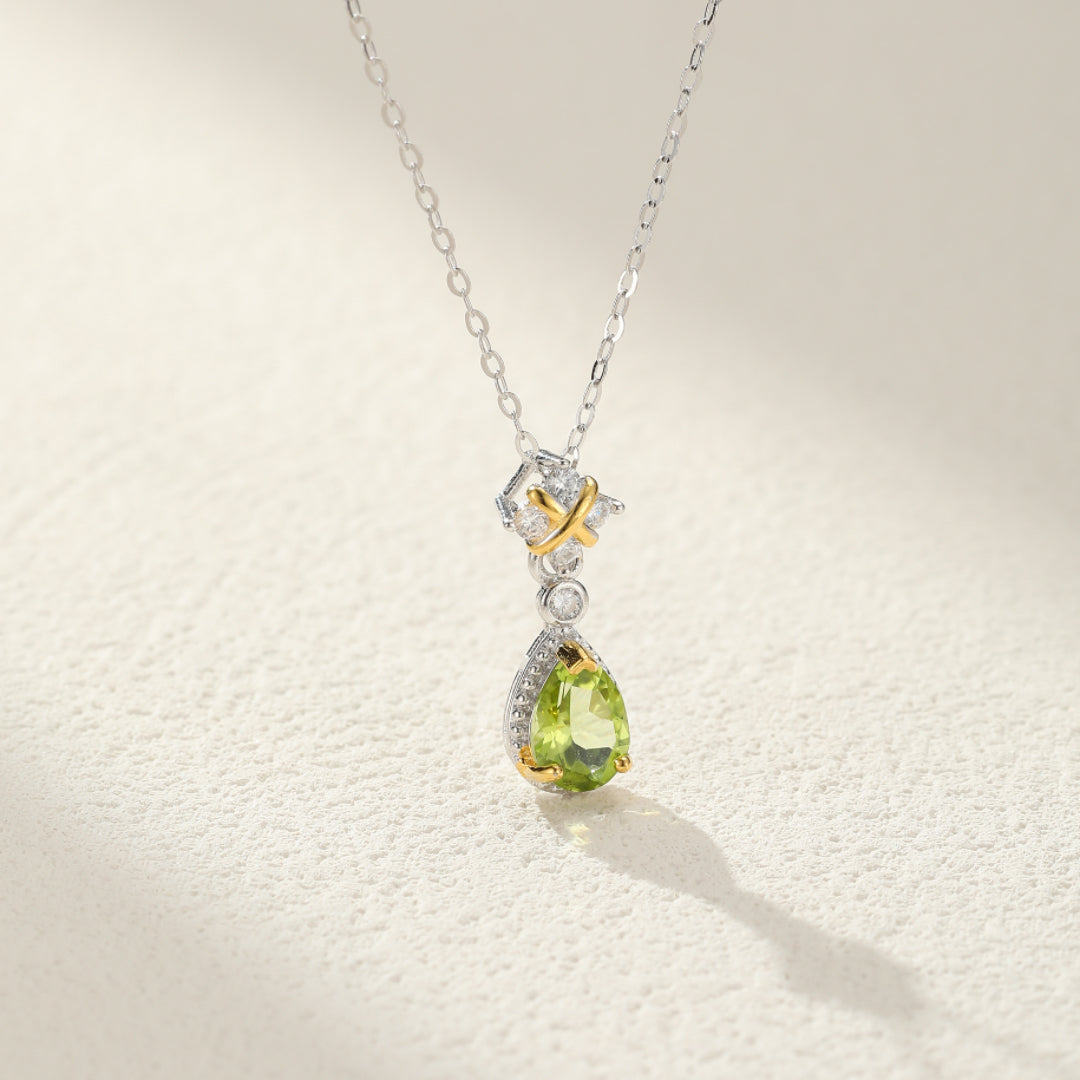 Un collier en argent avec une pierre de péridot en forme de goutte, orné de petits diamants et d'un détail doré en forme de X.