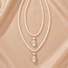 Deux colliers superposés en perles blanches, chacun orné d'un pendentif étoile dorée et d'un arbre de Noël stylisé avec une perle blanche à sa base, sur fond satiné beige.