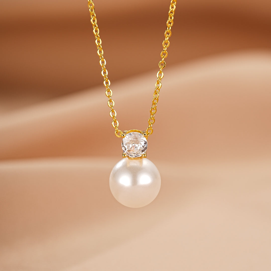 Collier en or jaune avec une perle blanche et un cristal brillant au centre, sur un fond beige satiné.