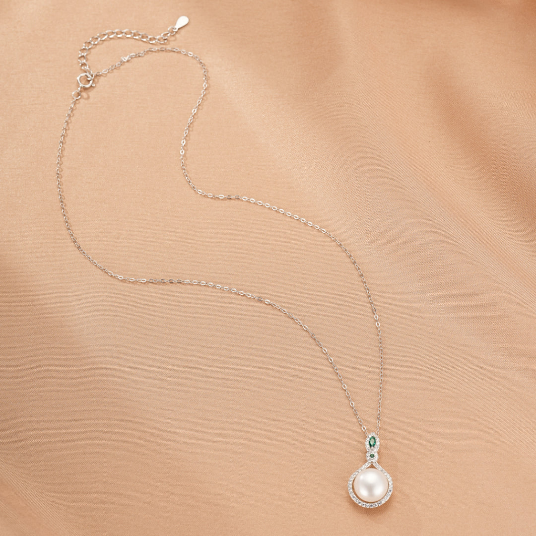 Collier en argent 925 avec une perle blanche centrale et des petites pierres vertes autour, sur un fond beige satiné.