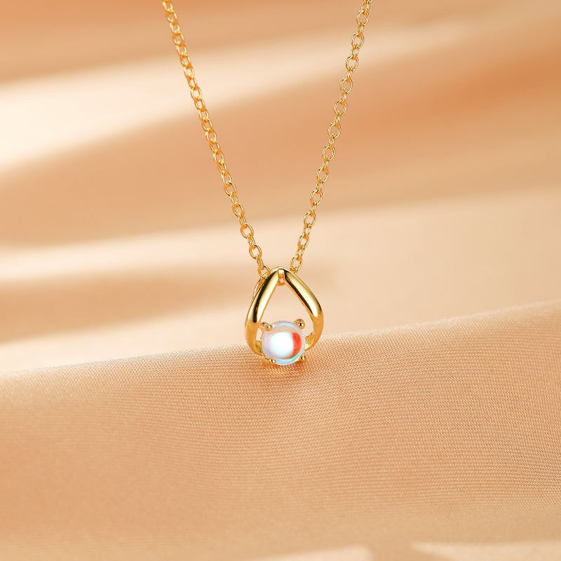 Collier en argent plaqué or avec un pendentif en forme de larme retenant une pierre de lune blanche aux reflets bleus et oranges, sur fond satiné beige.