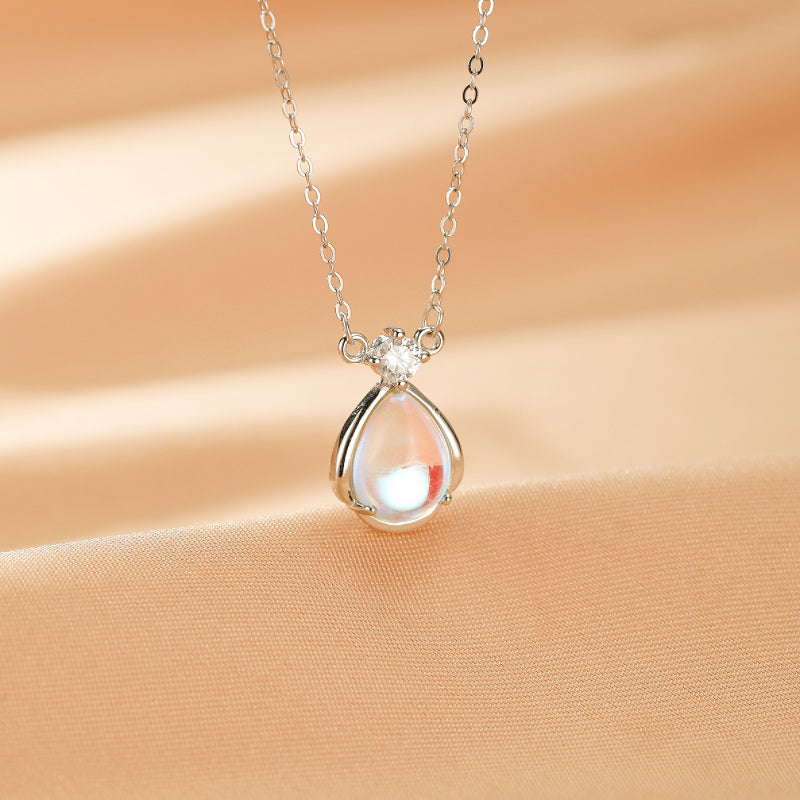 Collier argent "Larme Céleste" avec pendentif goutte de pierre de lune irisée et un petit diamant, sur fond satiné beige.