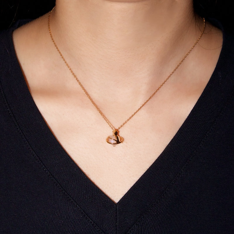 Collier plaqué or avec un pendentif "Orbite Céleste" en forme de sphère entourée d'anneaux, orné d'une pierre de lune lumineuse. Le collier est porté sur une peau claire sous un vêtement noir.