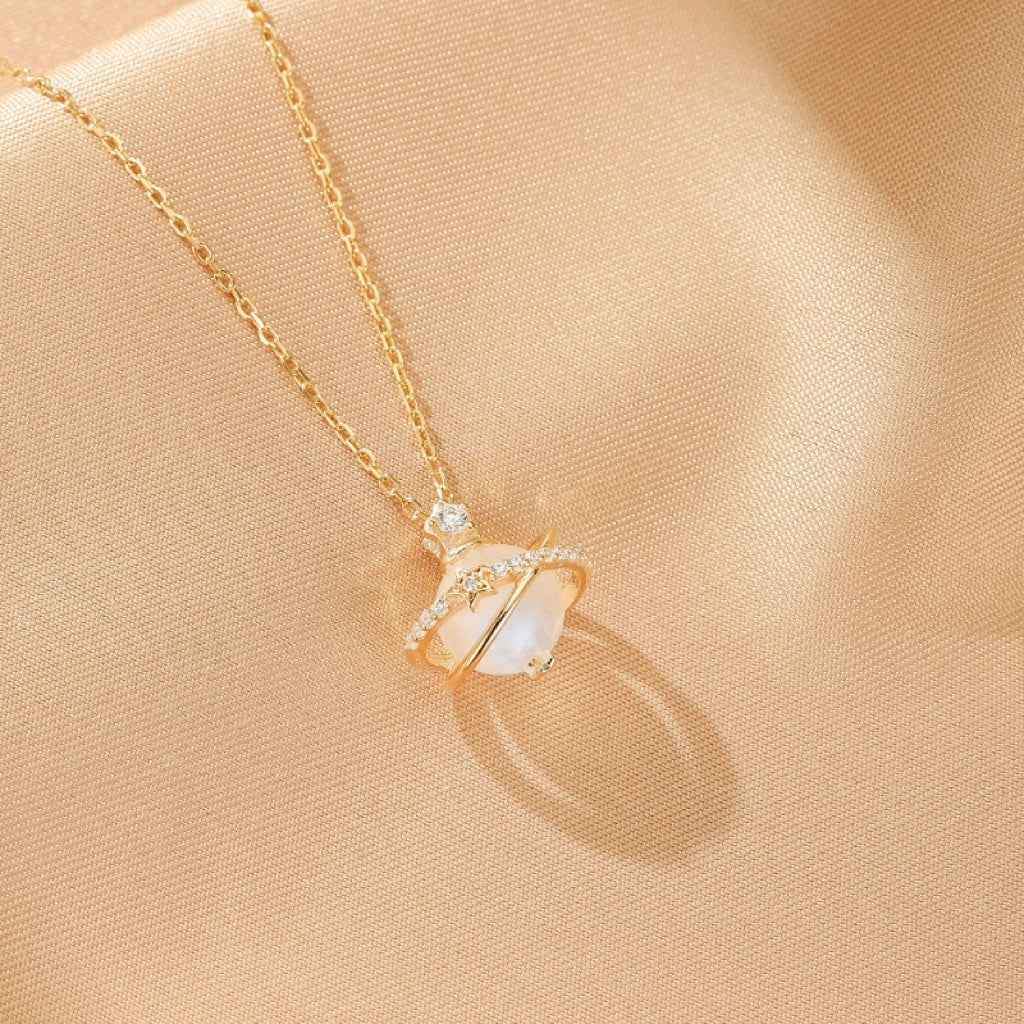 Collier plaqué or avec un pendentif en forme de planète, orné d'une pierre de lune blanche et d'une orbite de diamants, le tout sur un fond satiné.