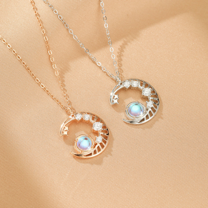 Deux colliers en argent, un plaqué or rose et l'autre argenté, avec un pendentif en forme de croissant de lune orné de petites étoiles et de cristaux, centré par une pierre de lune opalescente aux reflets bleus et roses.