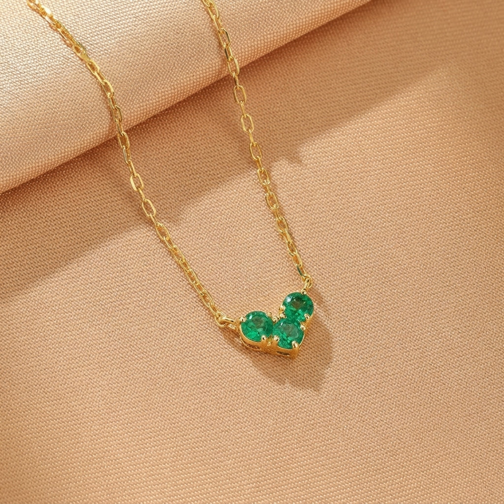 Collier en argent plaqué or avec trois pierres vertes disposées en forme de cœur, sur un fond beige