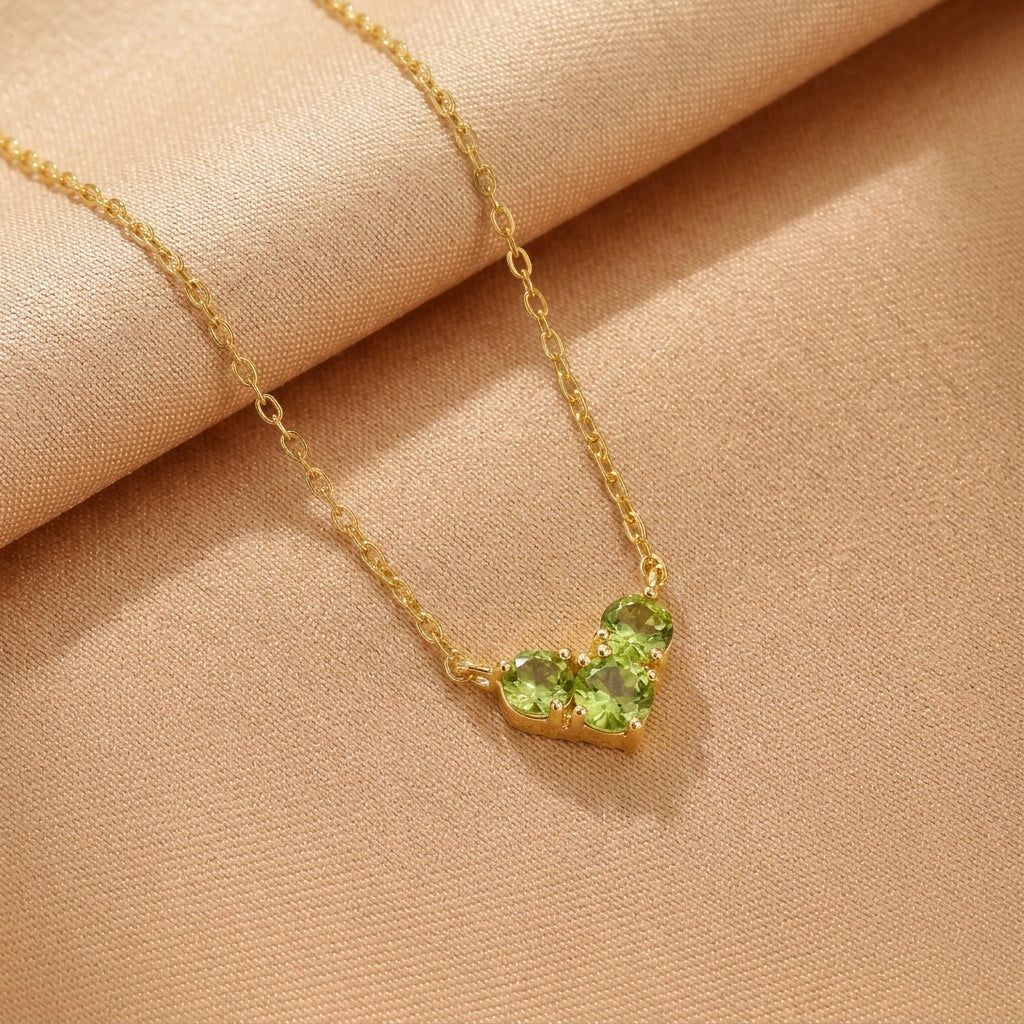 Collier en argent plaqué or avec trois pierres vertes disposées en forme de cœur, sur fond de tissu beige.