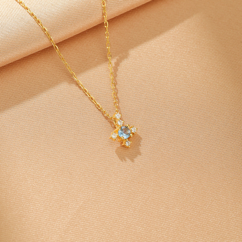 Collier en argent plaqué or avec une pierre bleue centrale entourée de petites pierres blanches, sur un fond beige texturé.