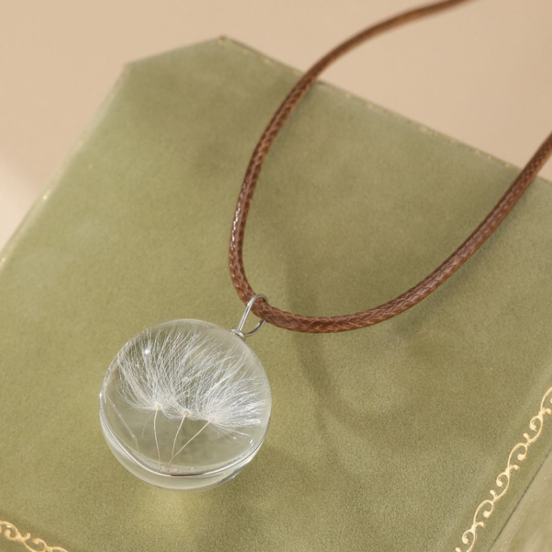 Collier avec pendentif sphérique transparent contenant des graines de pissenlit séchées, monté sur un cordon en cuir marron.