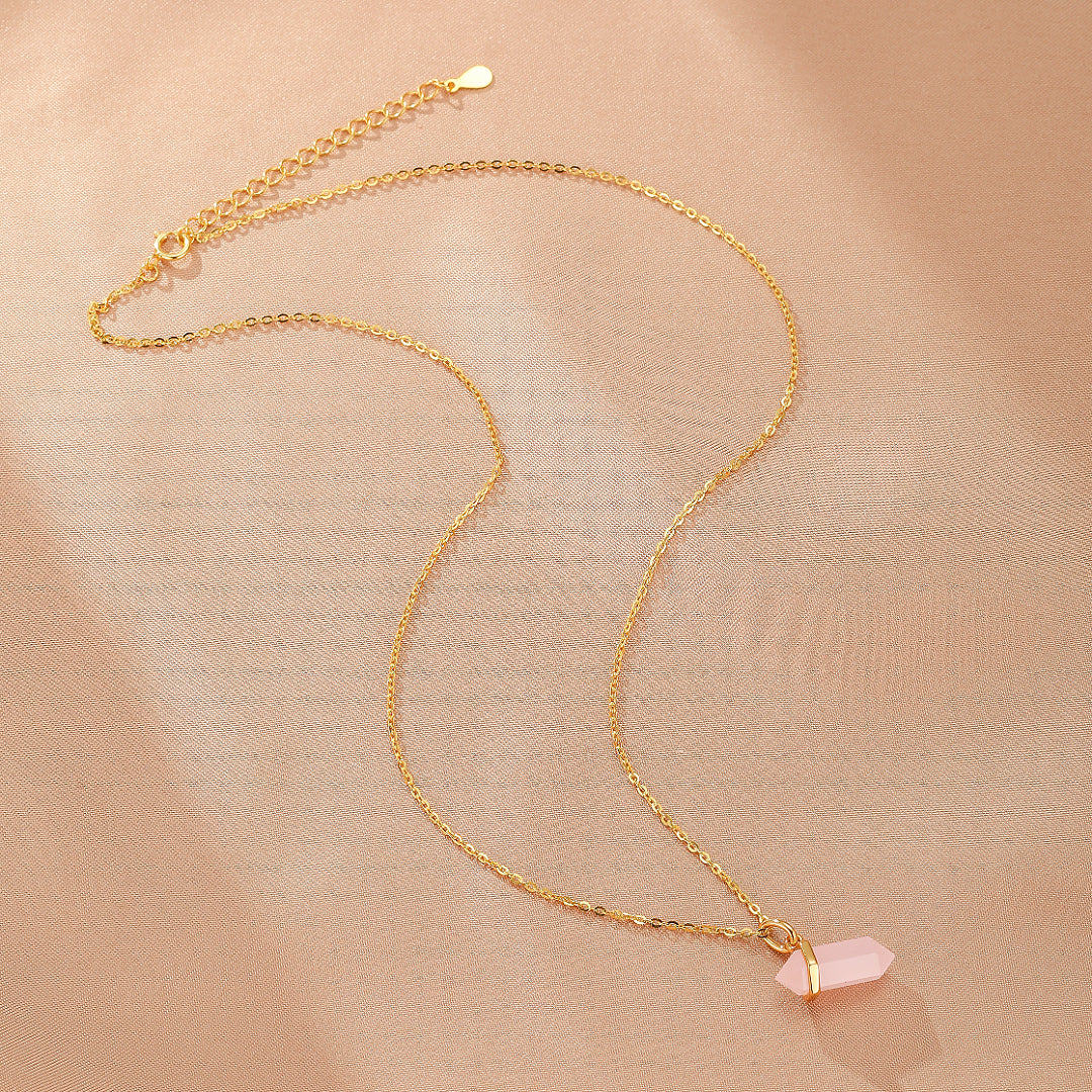 Collier fin en argent plaqué or 10K avec un pendentif en quartz rose brut taillé en pointe hexagonale, sur fond texturé rose pâle.