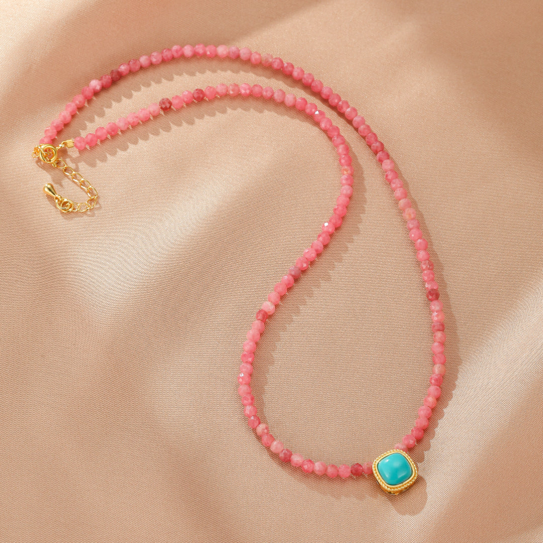 Collier fin en perles de rhodonite rose avec un pendentif carré en turquoise et fermoir doré réglable, sur un fond beige satiné.