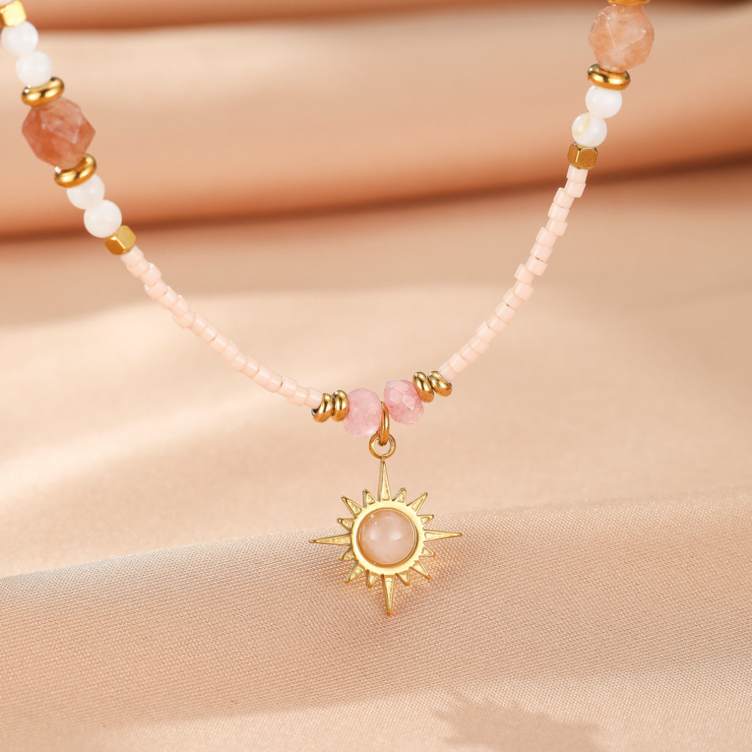 Gros plan d'un collier fait de perles roses pâles, de quelques perles dorées et de pierres naturelles roses, orné d'un pendentif en forme de soleil doré avec une pierre rose pâle au centre.