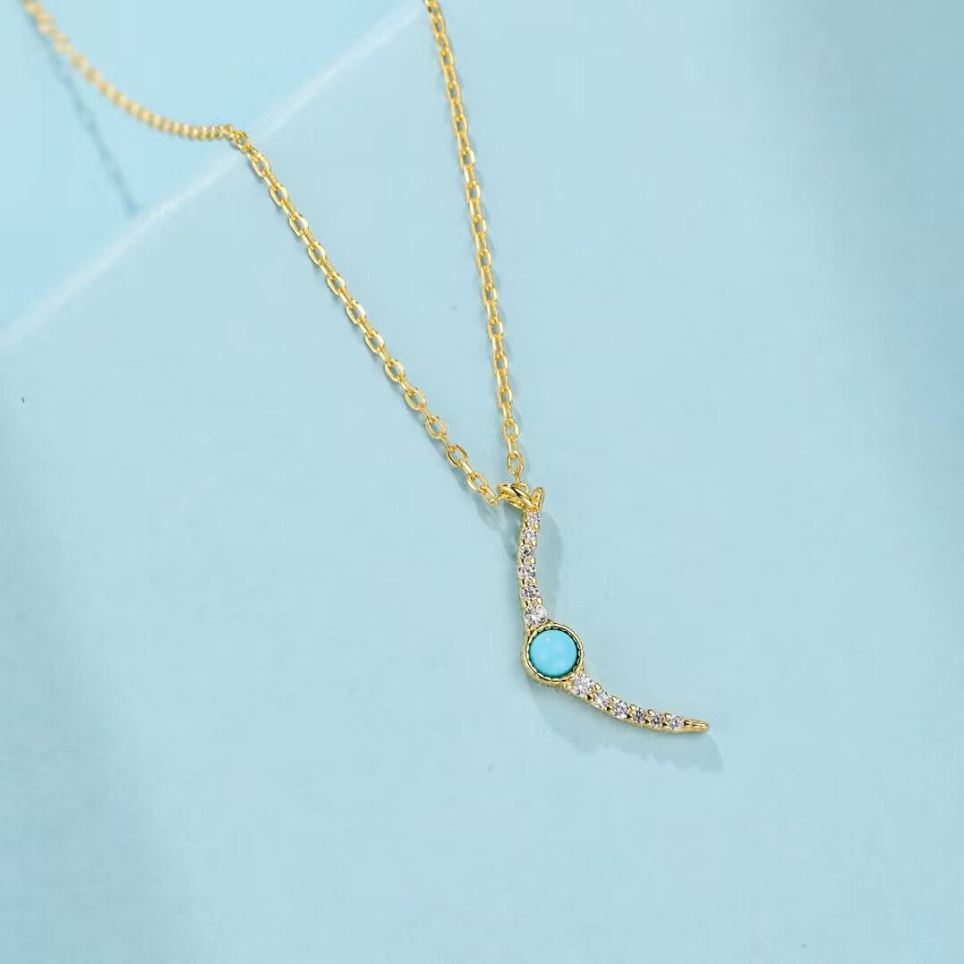 Collier en argent plaqué or avec une pierre turquoise ronde et des zircons cubiques incrustés formant un croissant de croissant de lune, sur fond bleu clair.