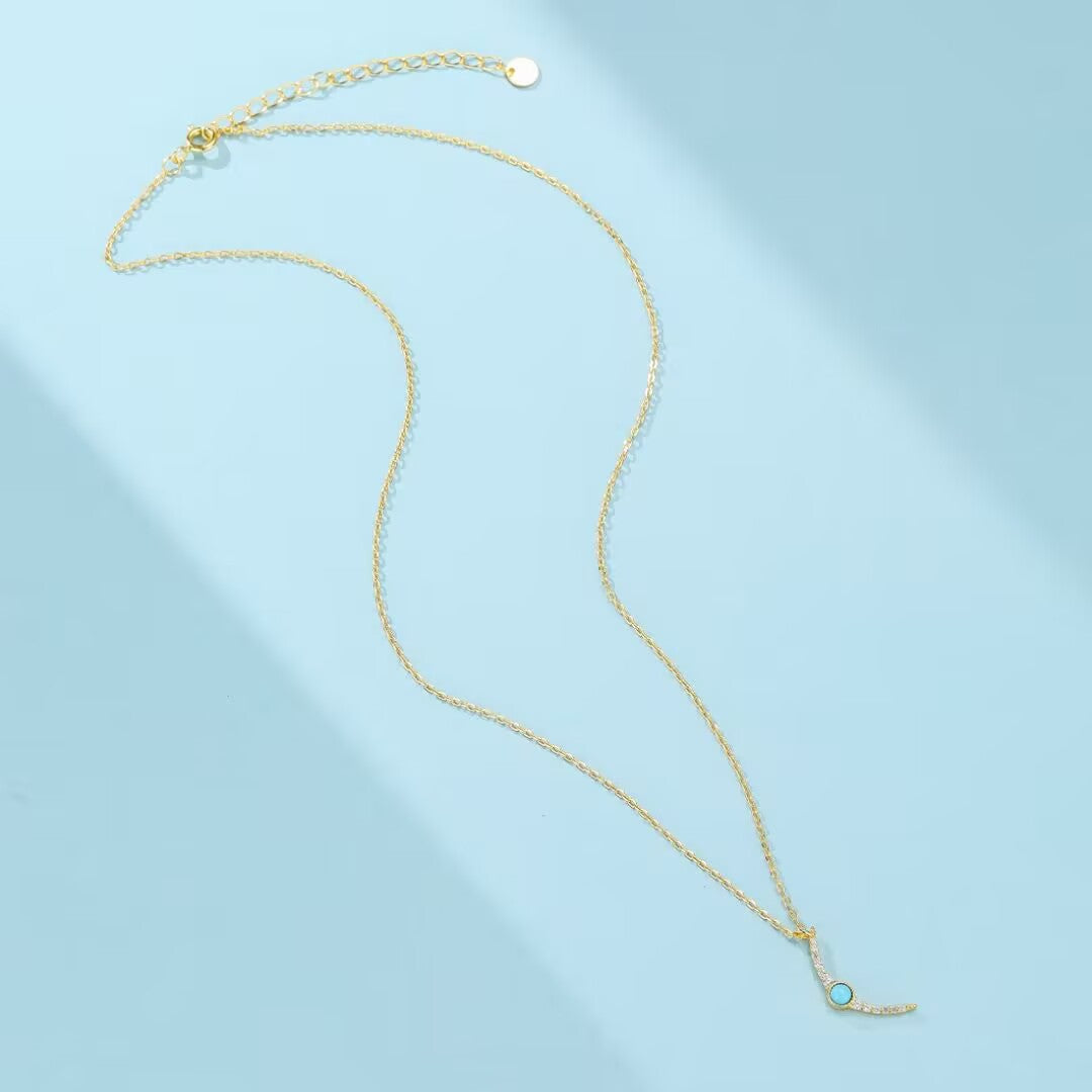 Un collier fin en argent plaqué or avec un pendentif en forme de L serti d'une petite turquoise ronde et de zircons diamants scintillants sur fond bleu clair.