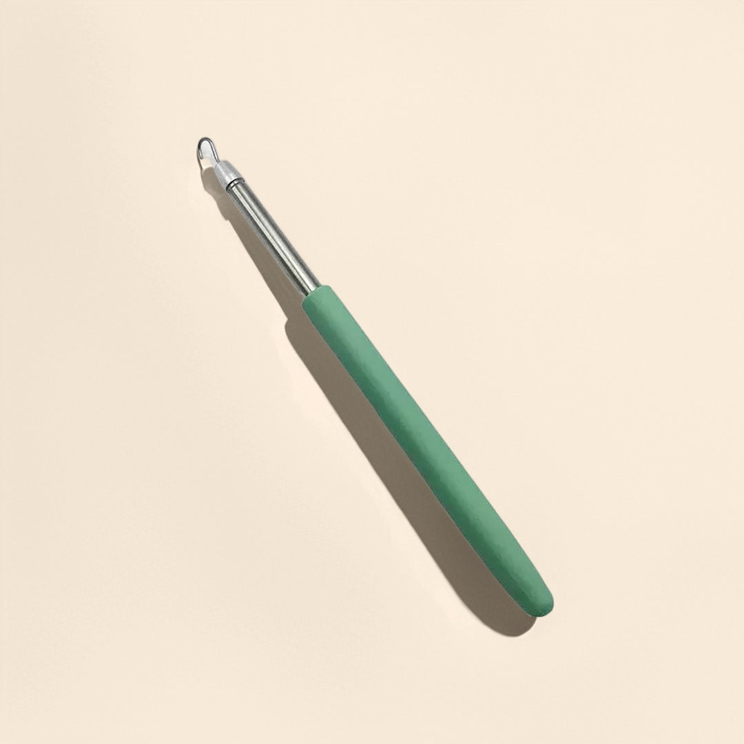 Crochet auxiliaire avec manche ergonomique vert et embout métallique fin, sur fond beige clair.