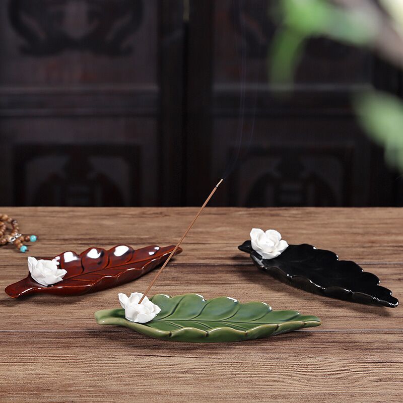 Trois porte-encens en forme de feuille de lotus en céramique, un vert, un marron et un noir, avec une fleur blanche décorative et un bâton d'encens allumé d'où s'échappe de la fumée.