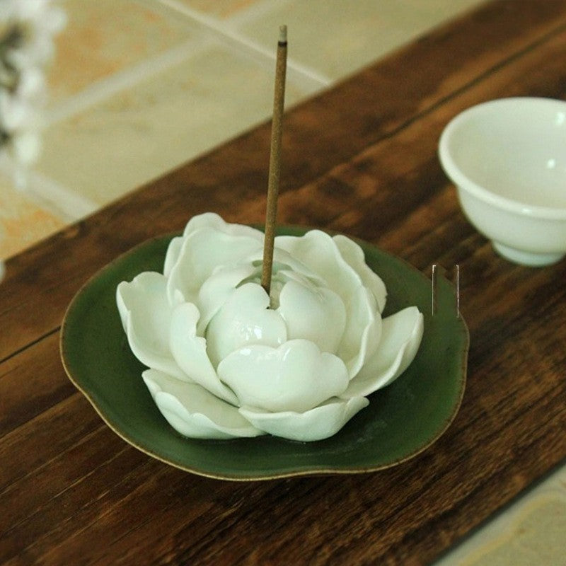 Encensoir en forme de pivoine blanche en céramique, avec un bâton d'encens allumé, posé sur une soucoupe verte, sur un fond en bois sombre.