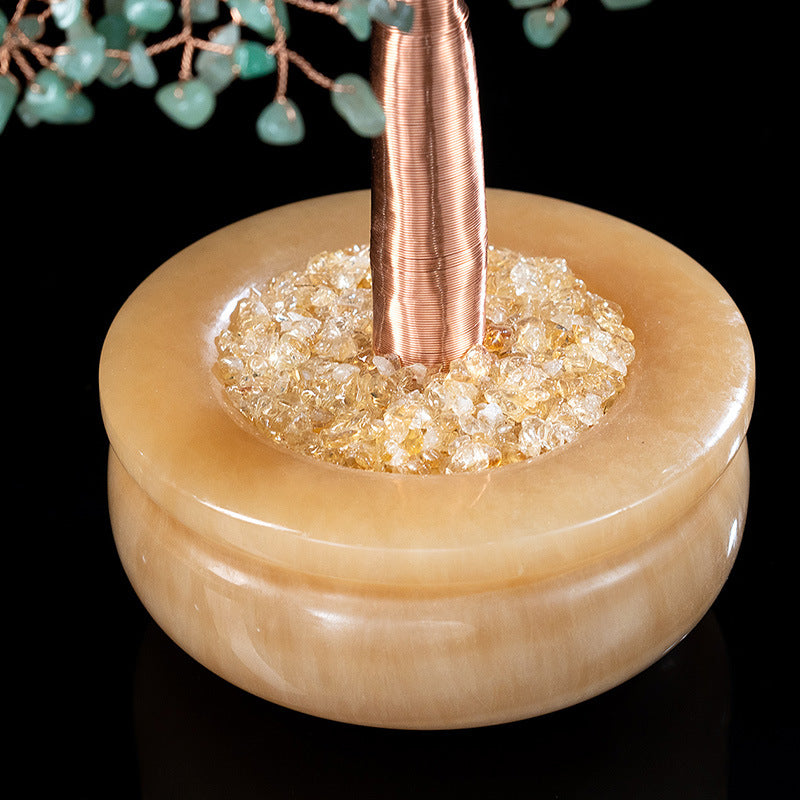 Gros plan sur la base d'un arbre de vie décoratif, composé d'une coupe en onyx clair remplie de citrine brute, avec un tronc en fil de cuivre et des feuilles d'aventurine verte visibles en arrière-plan.