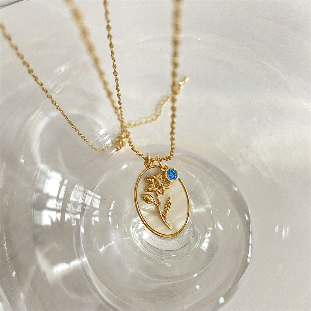 Pendentif ovale en nacre avec une fleur de jonquille dorée au centre, une petite pierre bleue sur le côté. Le pendentif est suspendu à une fine chaîne en or.
