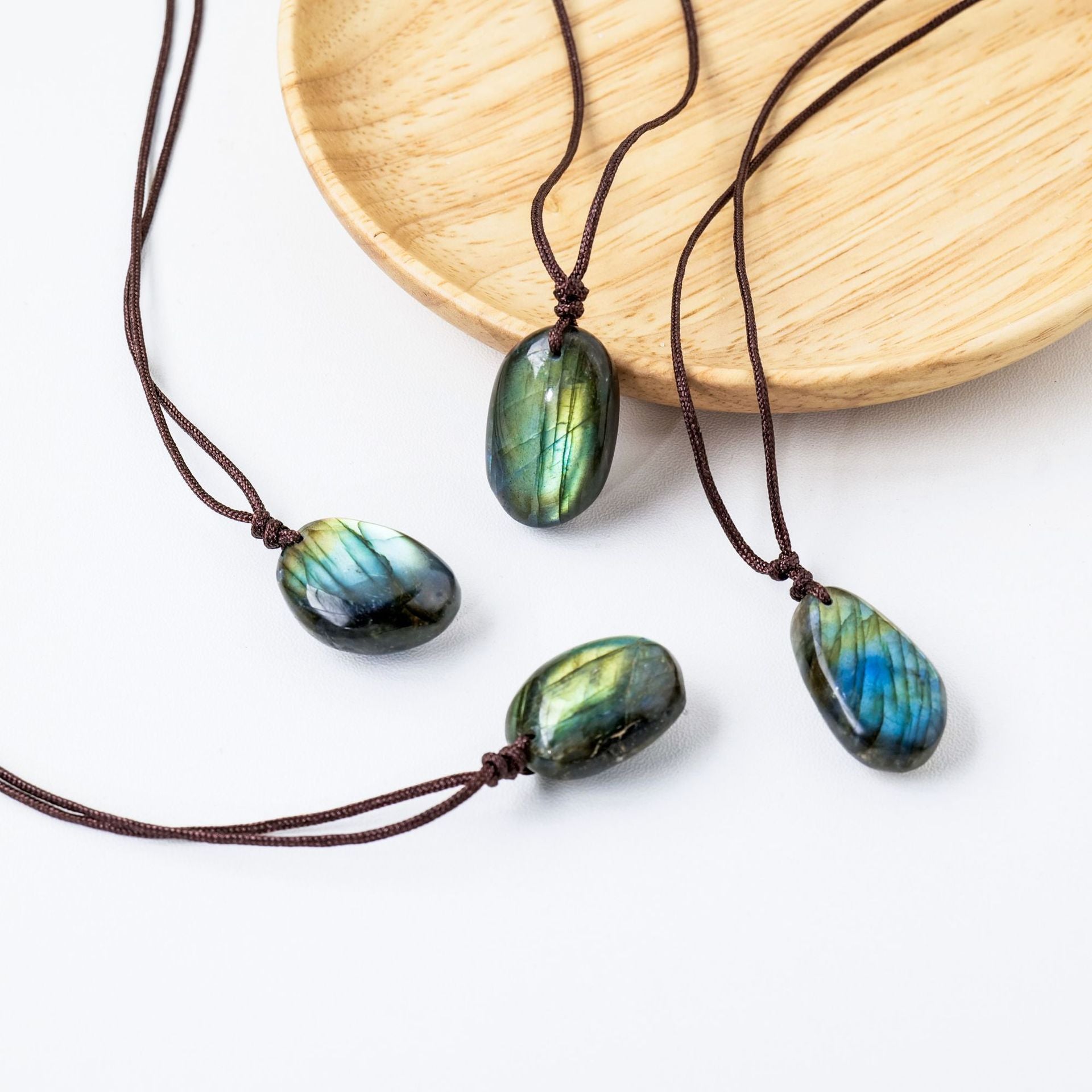 Quatre pendentifs en forme de galets de labradorite aux reflets bleus et verts, montés sur des cordons marron, disposés sur une assiette en bois et sur fond blanc.