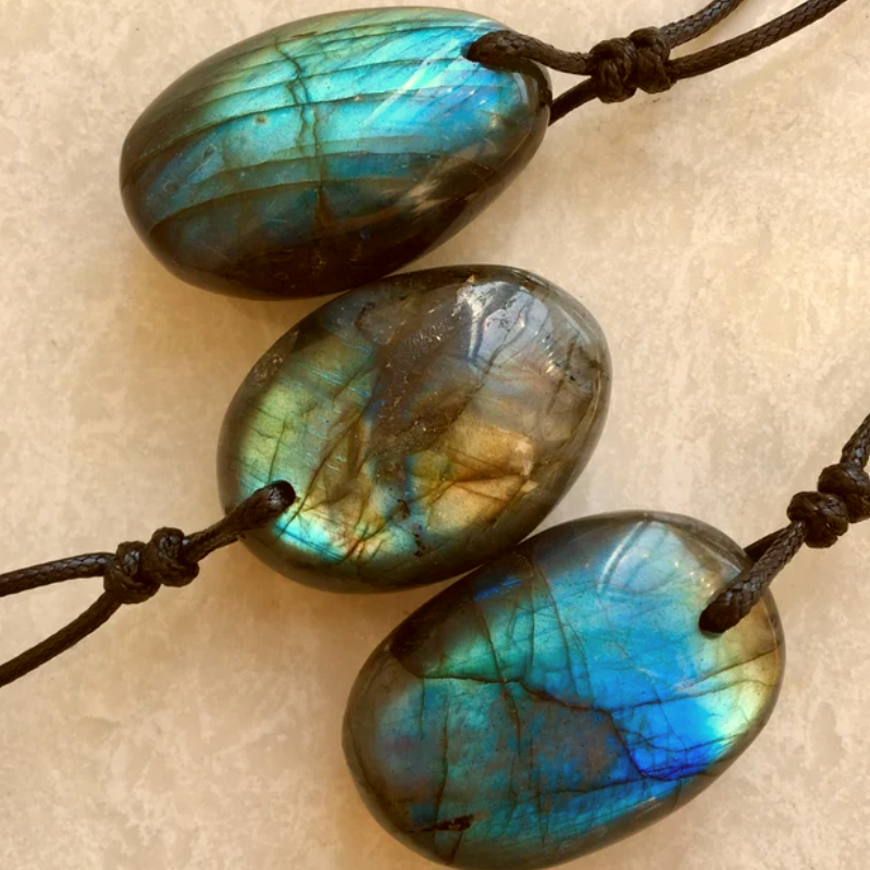 Trois pendentifs en forme d'œuf en pierre de labradorite aux reflets bleus et verts, chacun suspendu à un cordon noir.