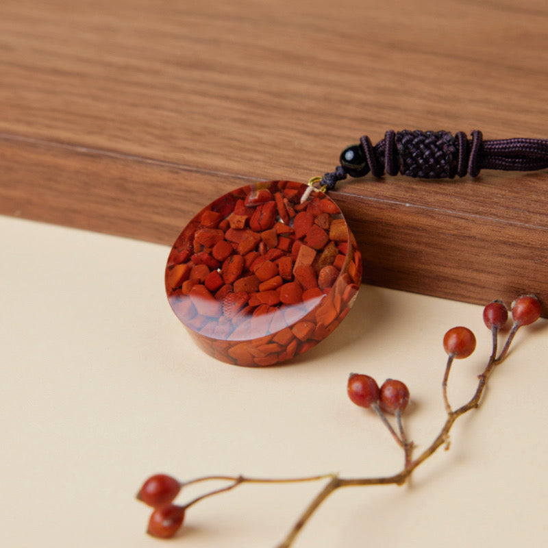 Pendentif rond en orgonite incrusté de petites pierres rouges, monté sur un cordon noir tressé, posé sur un fond en bois et un support clair avec quelques baies rouges.