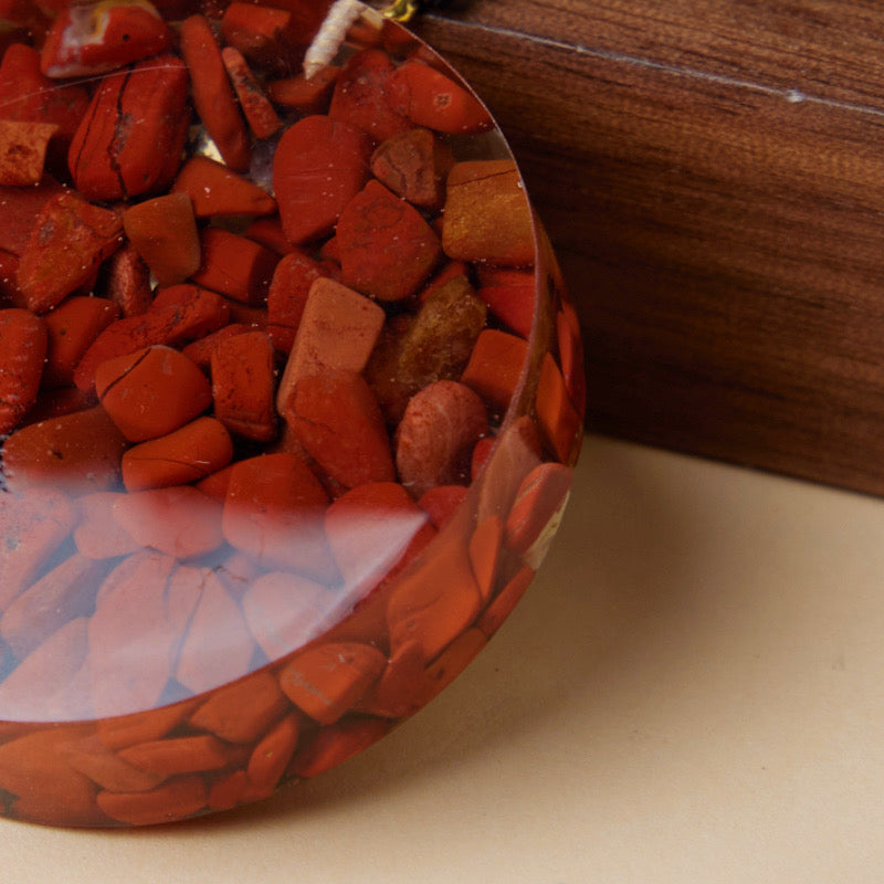Gros plan sur un pendentif orgonite aux éclats de jaspe rouge, incrusté dans une résine transparente et orné d'un motif œil mystique subtil.
