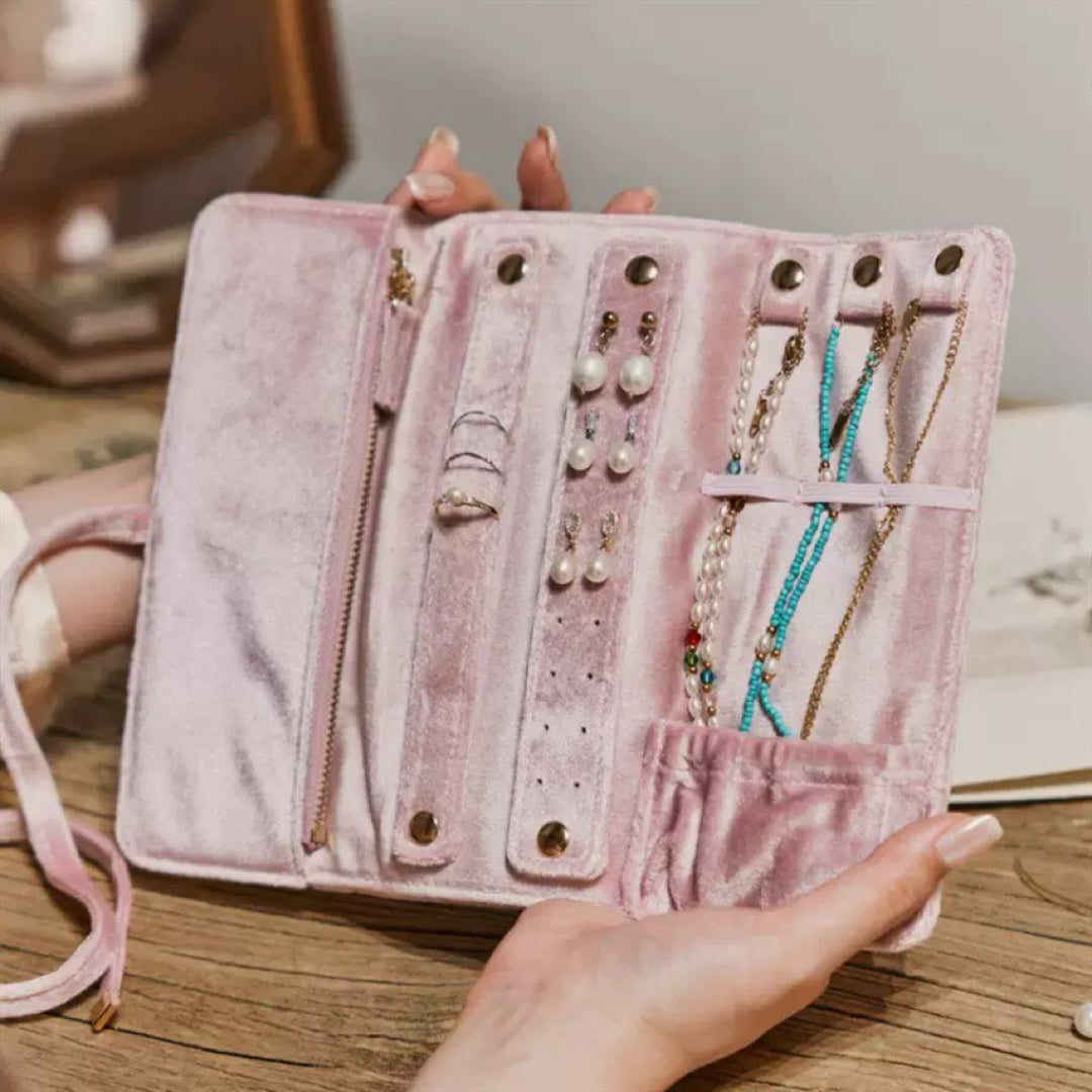 Pochette à bijoux rose en velours ouverte, montrant des colliers, des boucles d'oreilles et des bagues organisés, avec une main tenant la pochette.