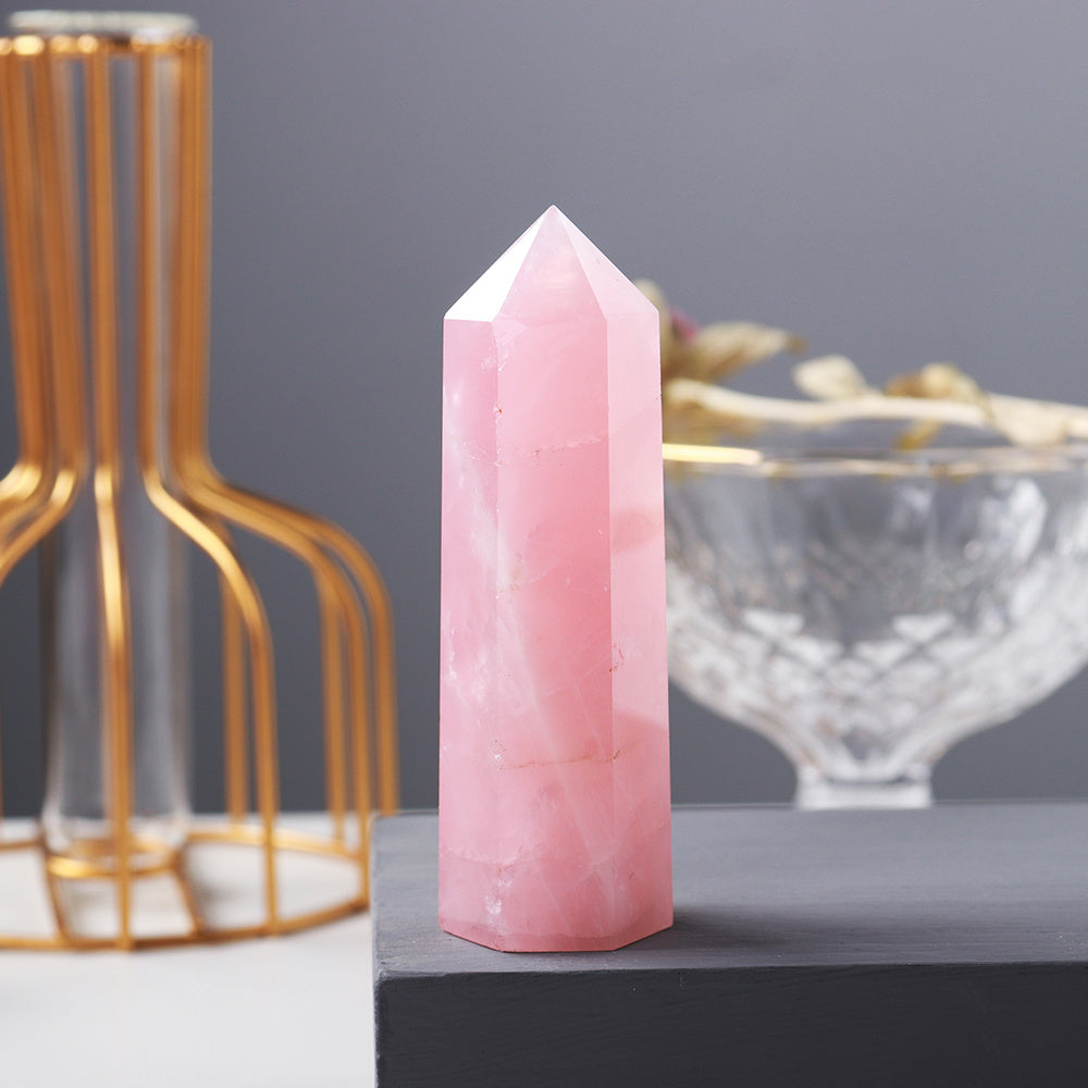 Pointe d'amour en quartz rose translucide avec des inclusions blanches, posée sur une surface grise, avec un vase doré et un bol en verre flous en arrière-plan.
